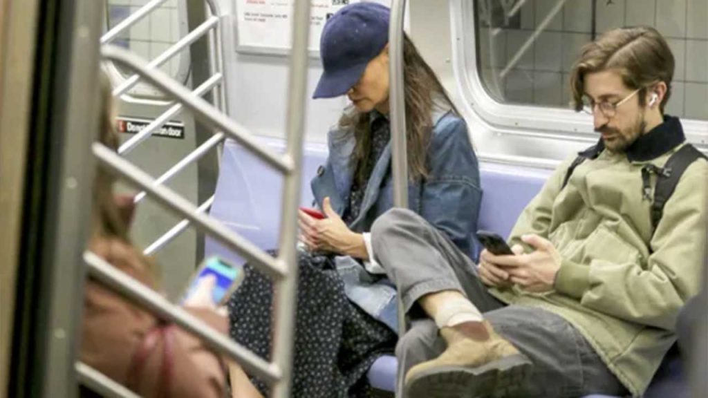 Kate Holmes sentada no metrô de boné mexendo no celular